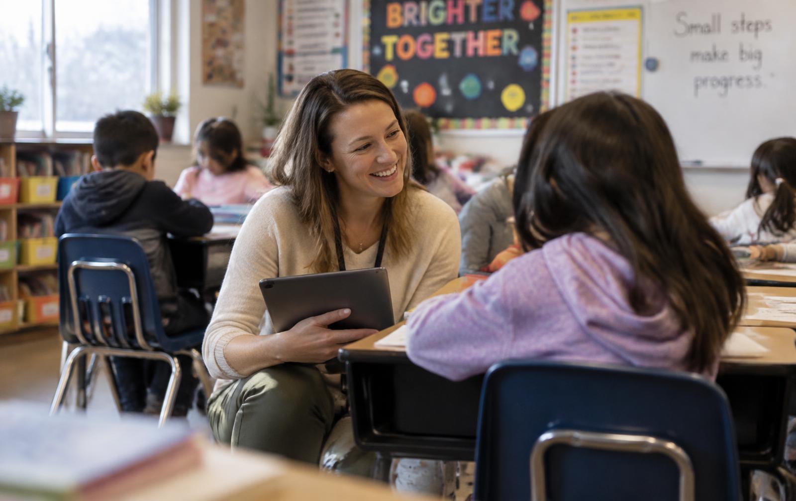 Teacher kneeling beside a student's desk with a tablet during a behavior check-in